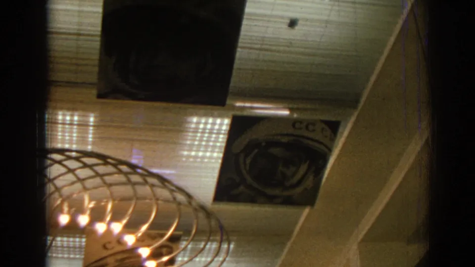 Izmir 1962 home-movie frame showing hanging astronaut portraits, partial Cyrillic-style lettering, and a ceiling light fixture.