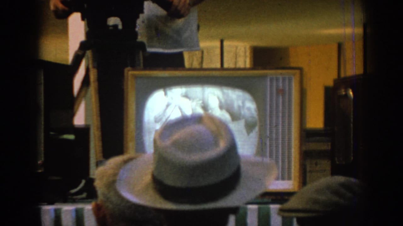 Adults in a dim room watch a small television set as a hat fills the foreground.