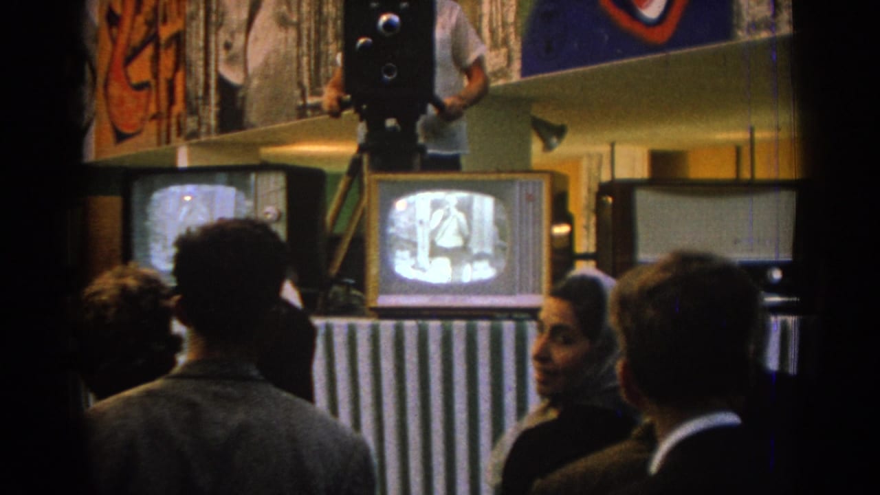 A group watches television while a man stands by a large camera on a tripod.