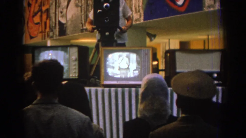 1962 Izmir home-movie frame showing adults from behind, including a woman with light hair and a man in a cap, watching televisions.