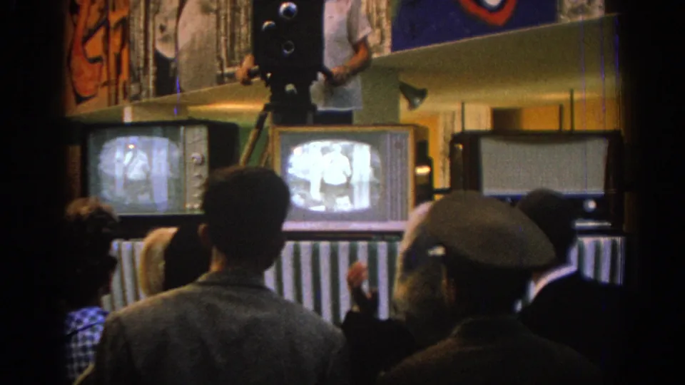 1962 Izmir frame of a crowded private room where adults watch bright television screens below a tripod-mounted camera.