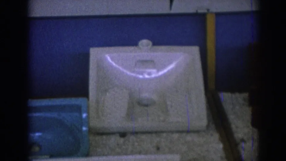Close 8mm view from 1962 Izmir of a white porcelain basin on a textured surface, with a cropped blue basin and wall panels behind it.