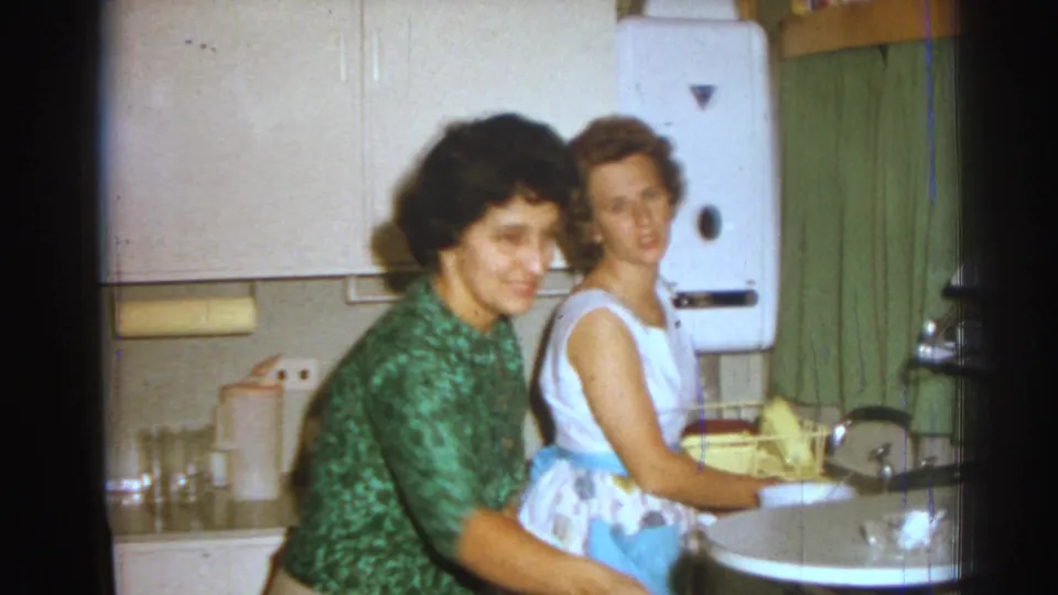 In a 1962 Izmir kitchen, two adult women stand by the sink, one in a green patterned top and one in a white dress with blue apron.