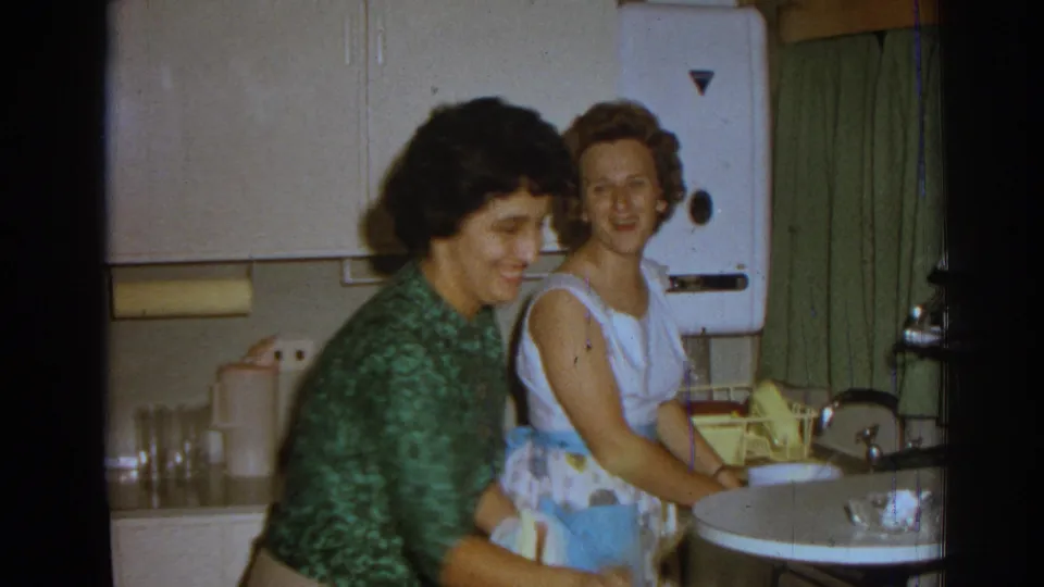 Two women in a 1962 Izmir kitchen smile near the sink, with a yellow dish rack, green curtains, and white wall appliance in view.