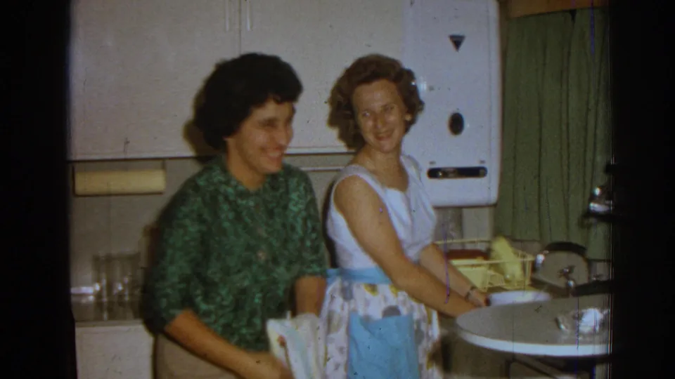 Two adult women laugh in a 1962 Izmir kitchen, framed with the sink, wall appliance, curtains, and household glassware.
