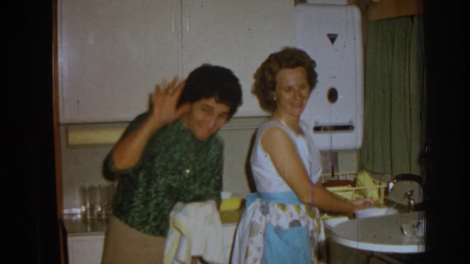 In the Izmir kitchen scene, one woman raises a hand toward the camera while another remains by the sink in a white dress and blue apron.