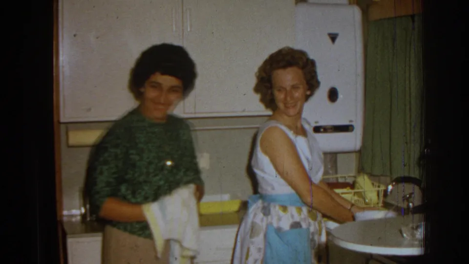 In a 1962 Izmir home interior, two women stand by a kitchen sink, one drying dishes while the other smiles toward the camera.