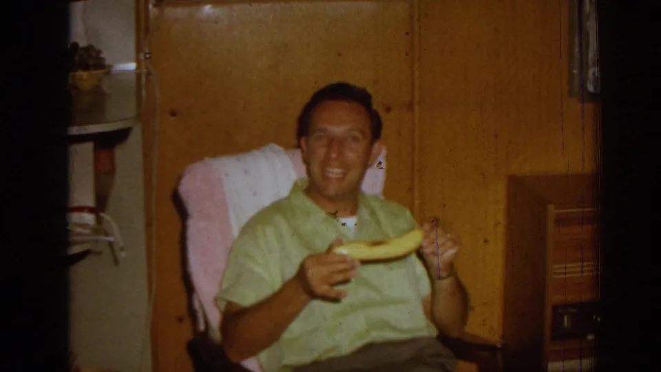 A man in a light green shirt sits in a wooden rocking chair in a 1962 Izmir home, smiling while holding a banana.