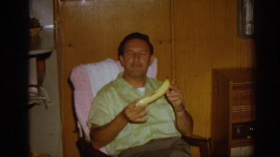 The seated man holds a banana while facing the camera in a private 1962 Izmir home interior with wood paneling.