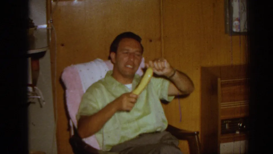 A man seated in a rocking chair gestures playfully with a banana in a 1962 Izmir home interior with wood-paneled walls.