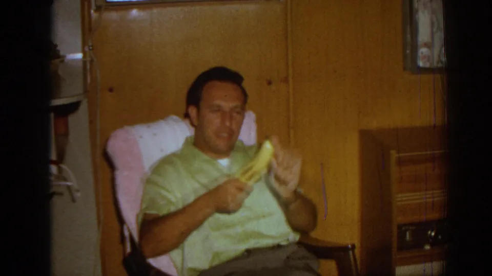 In 1962 Izmir home-movie footage, a man in a light green short-sleeve shirt sits in a wooden chair and gestures with a banana inside a wood-paneled private room.
