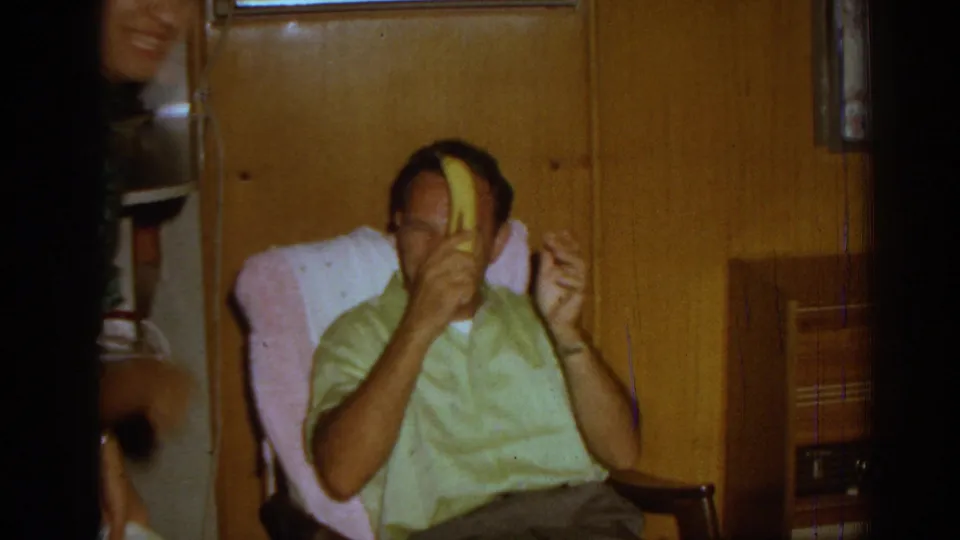 The man lifts the banana close to his face as if continuing the joke, seated against a wood-paneled private interior in the 1962 Izmir home movie.