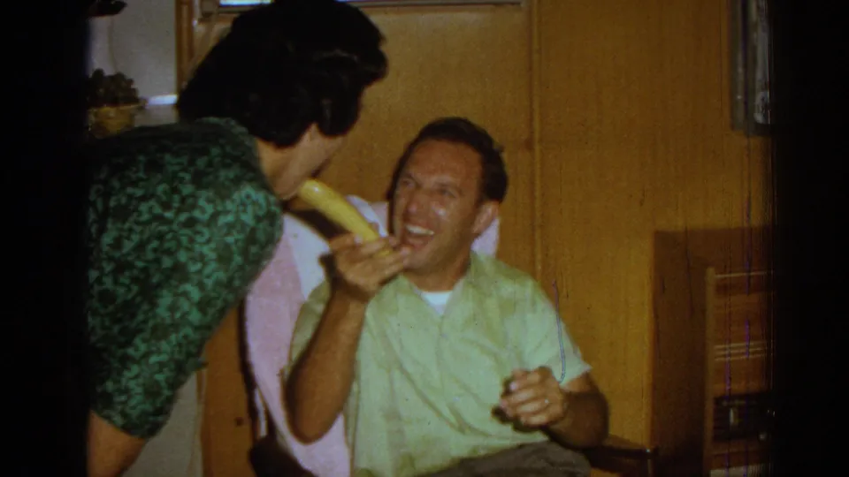 A woman in a green patterned dress leans toward the laughing seated man as he holds the banana, capturing the shared comic moment inside the Izmir private interior.