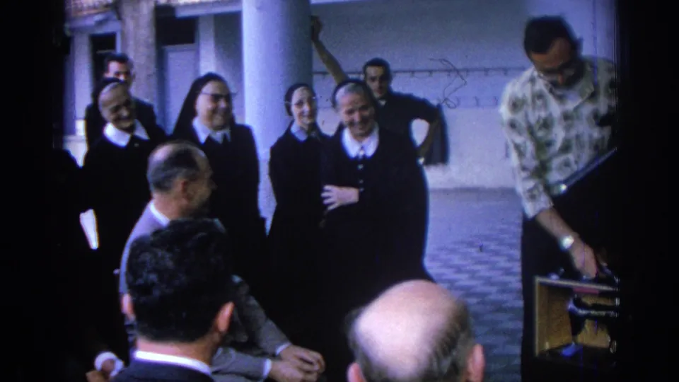 The Izmir convent courtyard group shifts position as nuns stand near visitors and a patterned-shirt man bends toward a chair.