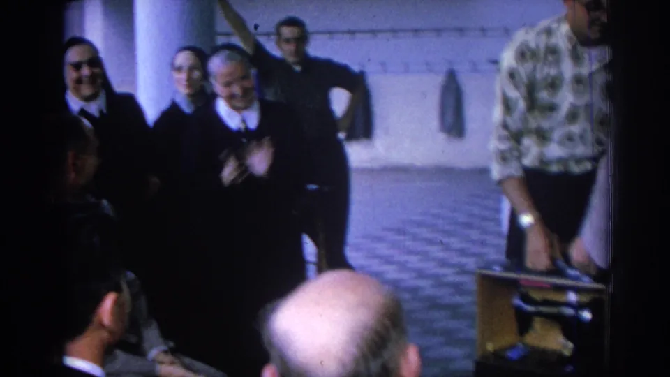 Late-frame view of the 1962 Izmir convent courtyard with smiling nuns, seated men, and a patterned-shirt visitor partly entering the side.