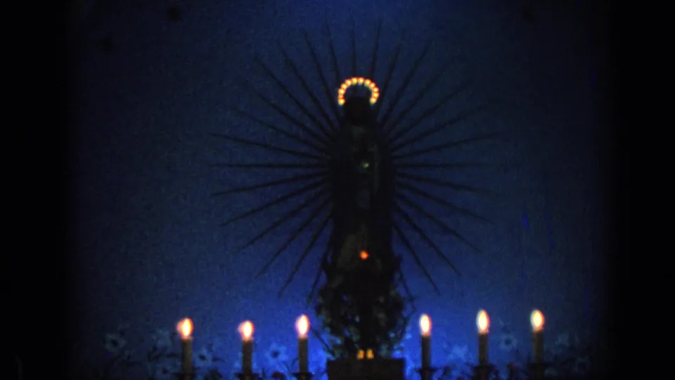 Low-light 1962 Izmir church frame with a haloed devotional statue, blue background, and multiple burning candles.