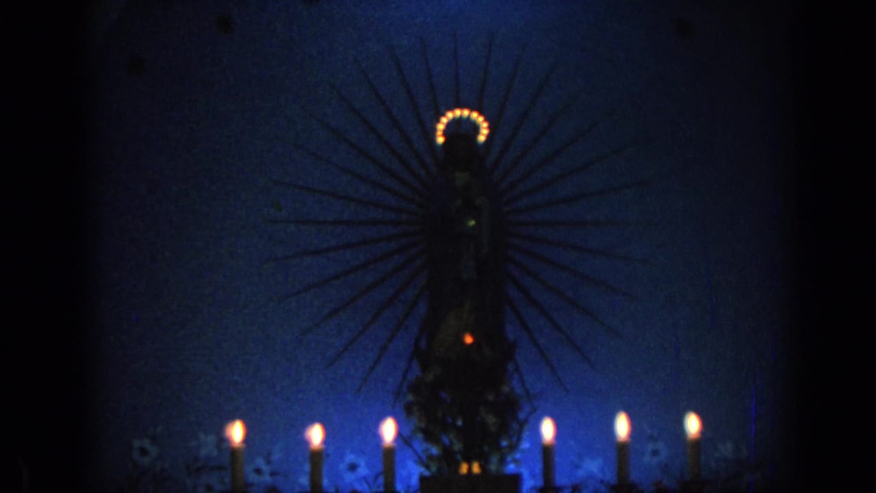 1962 Izmir 8mm frame showing a dark haloed Virgin Mary statue silhouette with lit candles below in blue nighttime light.