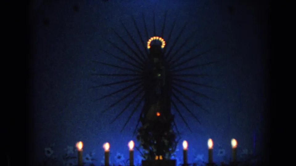 1962 Izmir 8mm frame showing a dark haloed Virgin Mary statue silhouette with lit candles below in blue nighttime light.