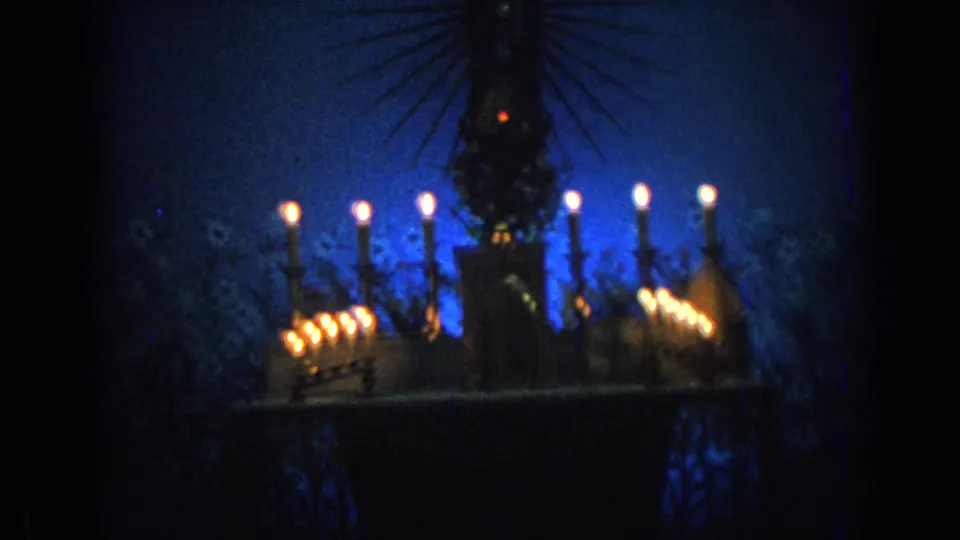 1962 Izmir 8mm frame of the candlelit Virgin Mary statue display, with flames lining a dark shrine under blue light.