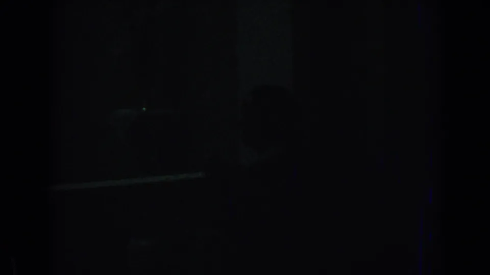 Underexposed home-movie frame showing a dark private interior with a dim rectangular form and little identifiable detail.