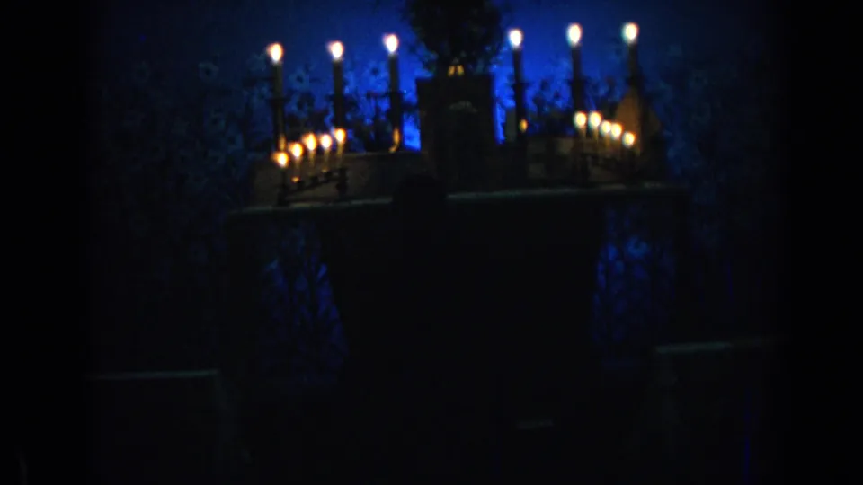 1962 Izmir home-movie frame showing lit candles on a shelf or mantel against blue illumination in a dark private interior.