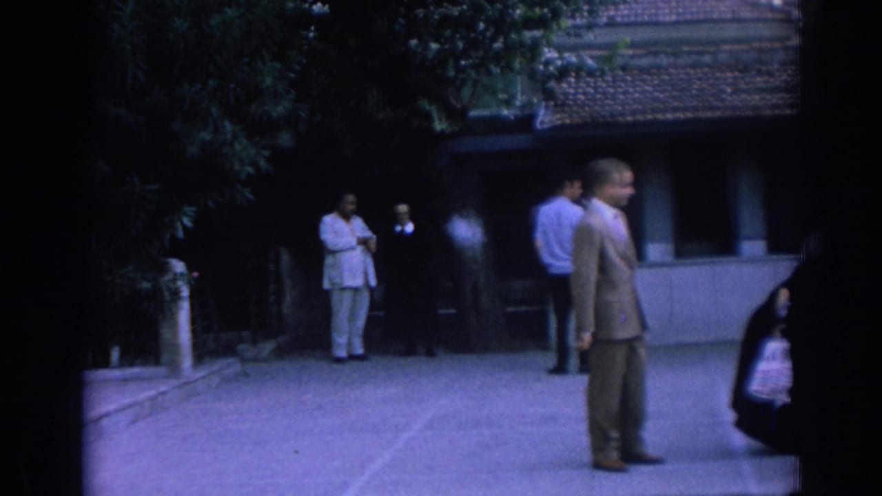 Adults stand in a shaded paved courtyard near trees and a tiled-roof building.