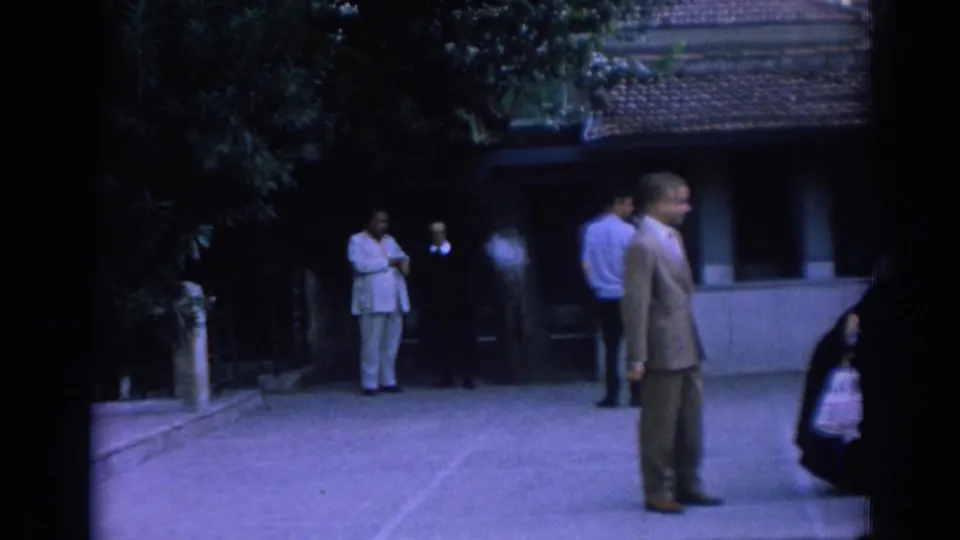 1962 Izmir home-movie frame showing adults in a shaded courtyard near leafy trees and a tiled-roof structure.