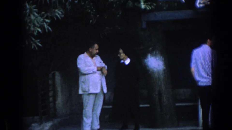 1962 Izmir frame of a man in a light suit and a woman in dark clothing standing near trees and a wall.
