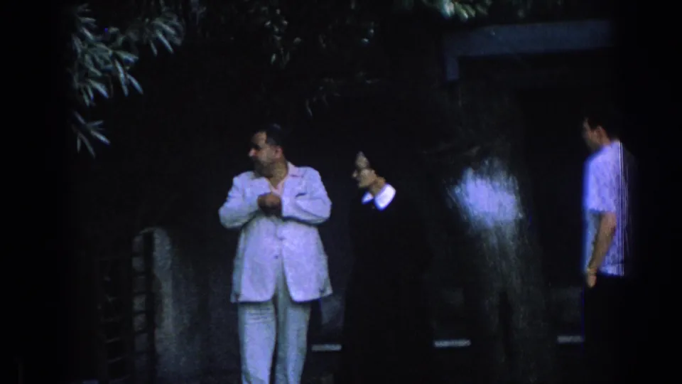 1962 Izmir home-movie frame with a suited man, a woman in dark clothing, and a passing young man in a courtyard.