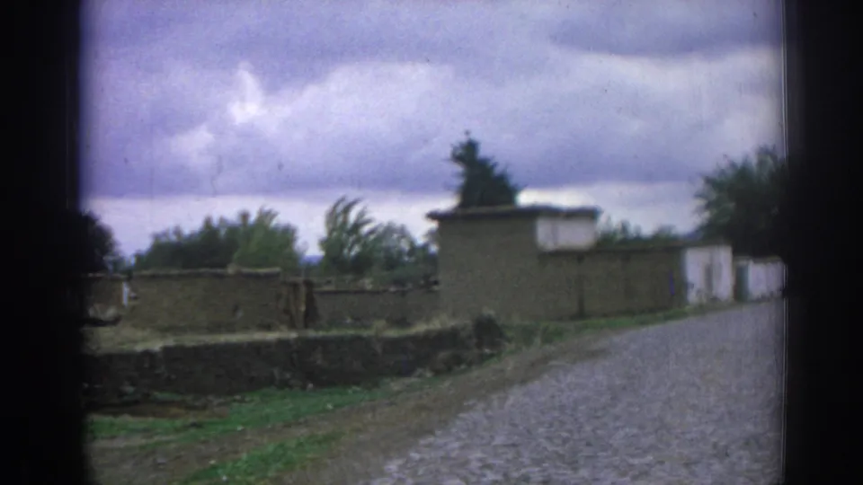1962 Izmir home-movie frame showing a cobblestone road, low walls, earth-toned buildings, trees, and overcast sky.