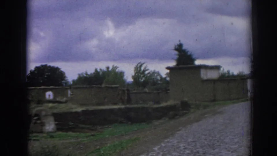 1962 Izmir frame with low stone walls, trees, earth-toned buildings, and a cobblestone road curving through the neighborhood.