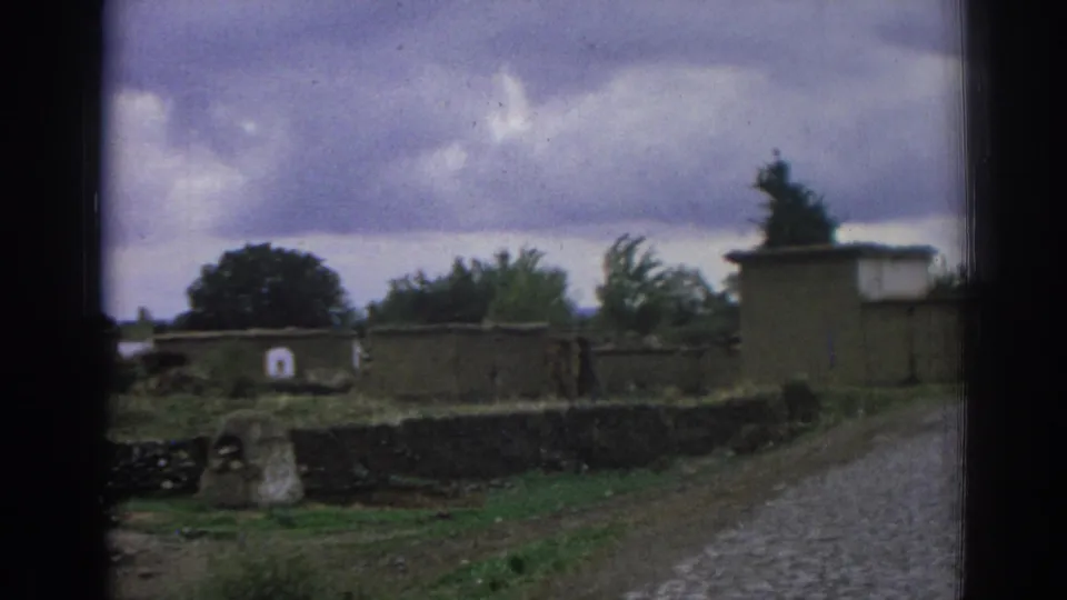 1962 Izmir home-movie frame showing a small stone structure, low walls, trees, and the edge of a cobblestone road.