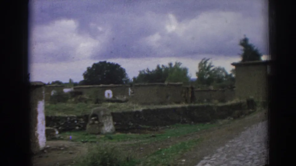 1962 Izmir frame with a small stone structure in front of low walls, greenery, and a cobblestone road under heavy clouds.
