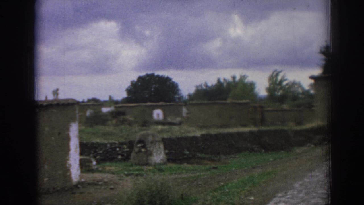 1962 8mm view in Izmir, Turkey, showing a quiet cemetery area with stone walls, low markers, grass, a dirt path, and overcast sky.