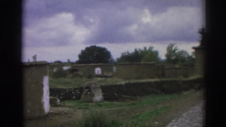 1962 8mm view in Izmir, Turkey, showing a quiet cemetery area with stone walls, low markers, grass, a dirt path, and overcast sky.