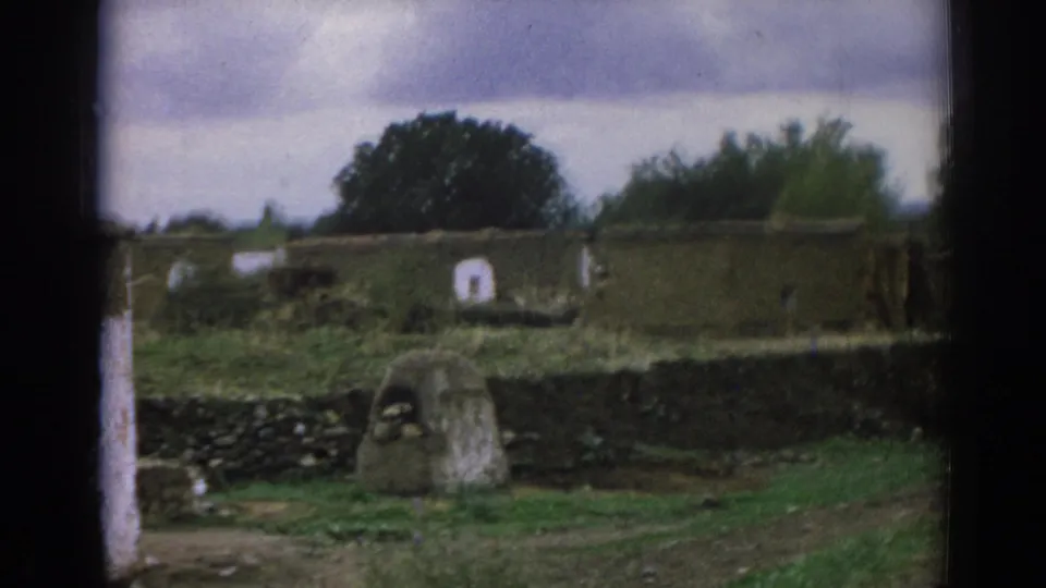 1962 Izmir cemetery view with a central low stone grave marker, stacked stone wall, grass, whitewashed features, trees, and gray sky.