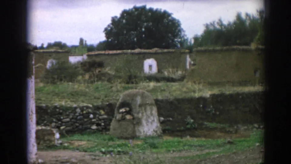 Closer 1962 Izmir cemetery frame showing a rounded stone grave marker before a dark stone wall, with whitewashed masonry, grass, and trees beyond.