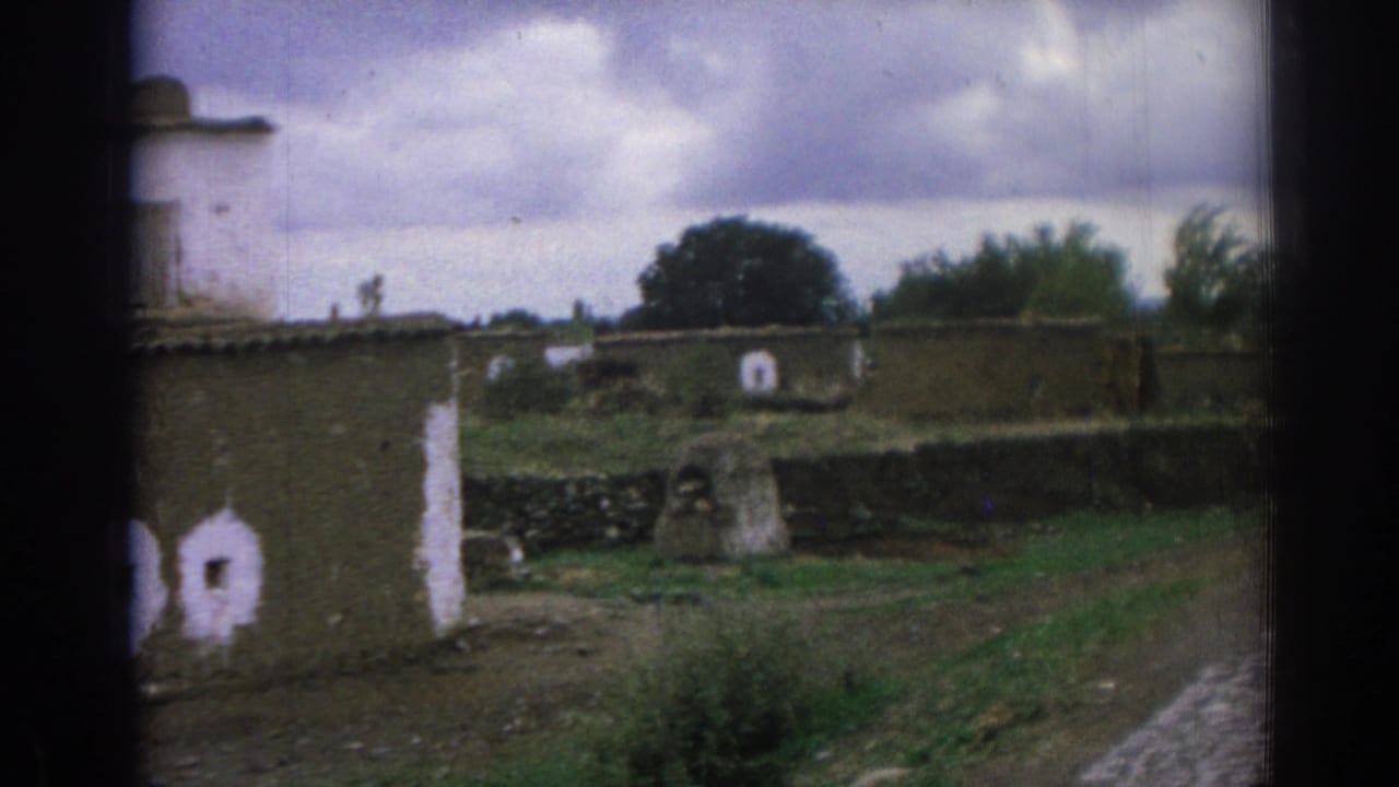 1962 Izmir-area 8mm frame showing weathered rural homes, white-painted openings, grass, and an unpaved path beneath overcast clouds.