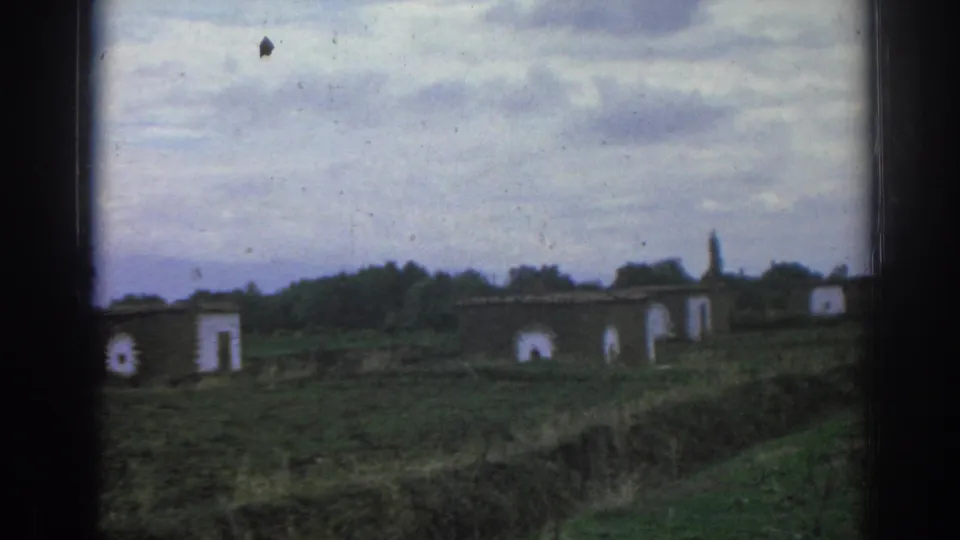 1962 Izmir-area 8mm frame showing a wider rural view with low buildings, white wall details, open ground, trees, and heavy clouds.