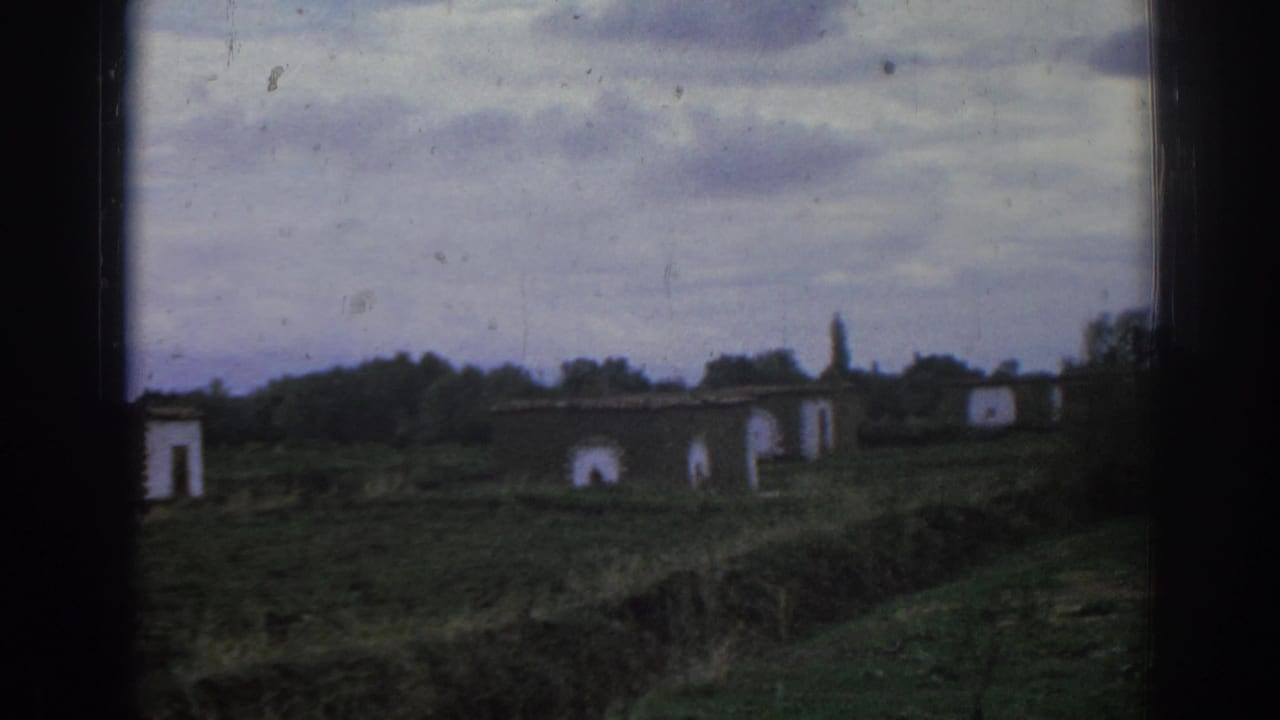 Distant abandoned buildings with arched openings in a grassy field under clouds.
