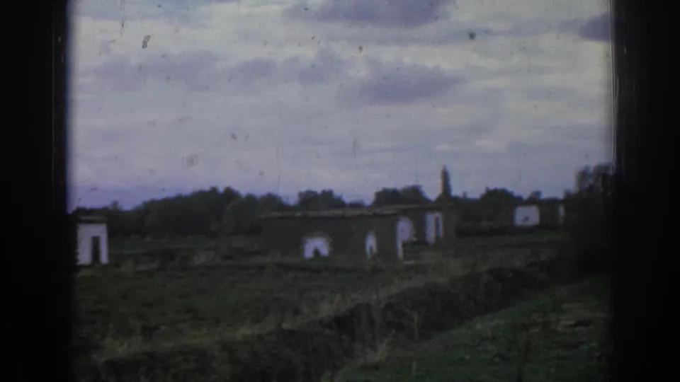 1962 Izmir home-movie frame showing weathered rural buildings with arched openings across a grassy, deserted field.