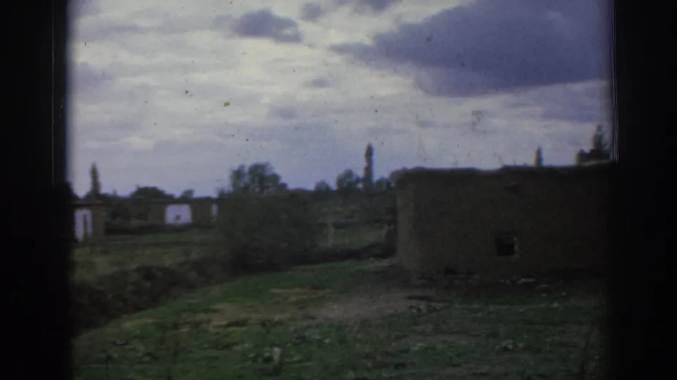 1962 Izmir, Turkey home-movie frame showing a low earthen building, fields, and cloudy sky in a damaged rural setting.