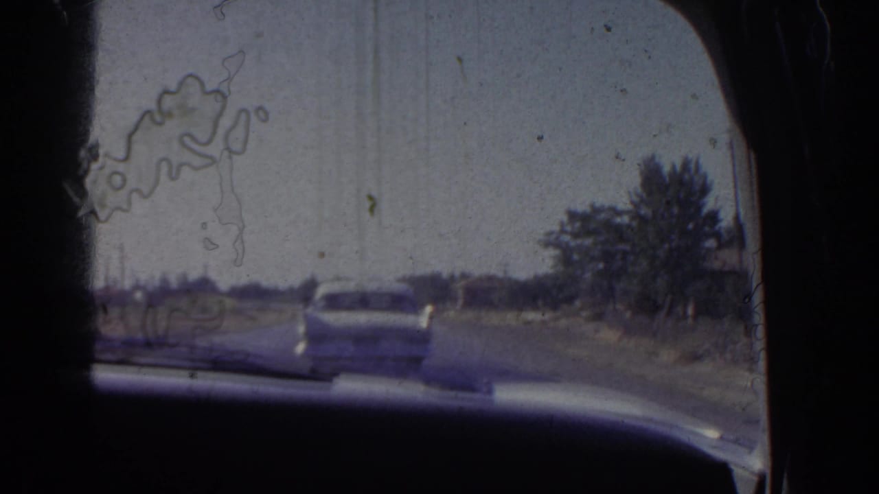 Dashboard-framed view through a windshield following a white sedan on a rural road.