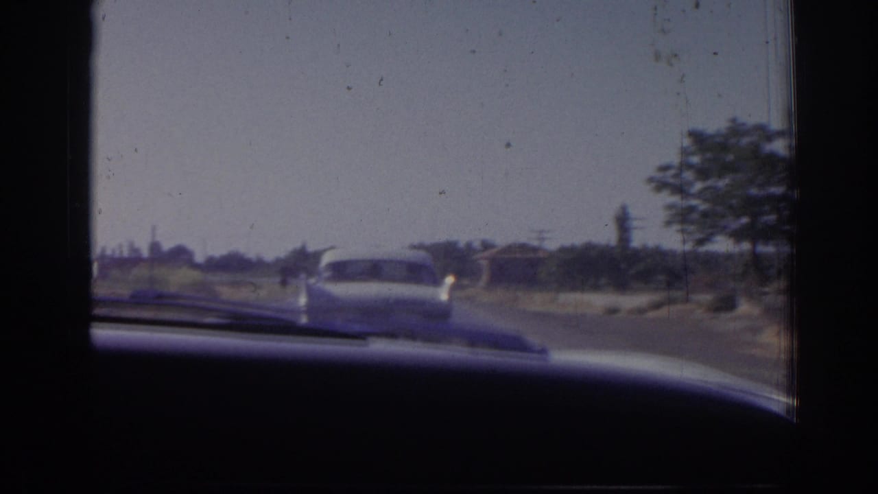 White sedan ahead on a paved road, seen past the dashboard and windshield frame.