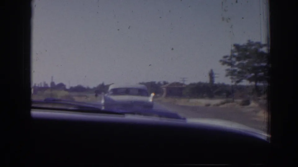Passenger-side windshield view in 1962 Izmir footage, with a white sedan ahead and low roadside structures in the distance.