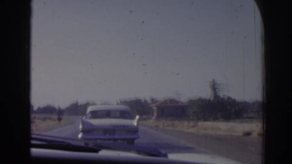 1962 Izmir road-travel frame showing a white sedan ahead, viewed through a windshield beside sparse trees and a low building.