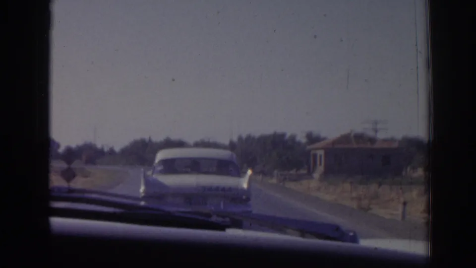 Late frame from the 1962 Izmir driving clip, with a sedan centered ahead and a low building along the right side of the road.