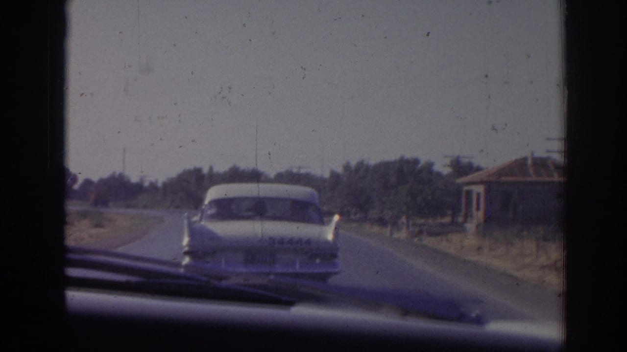 1962 Izmir home-movie view from inside a moving car following a white sedan on a rural road with trees and a low building nearby.