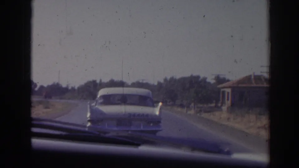 1962 Izmir home-movie view from inside a moving car following a white sedan on a rural road with trees and a low building nearby.