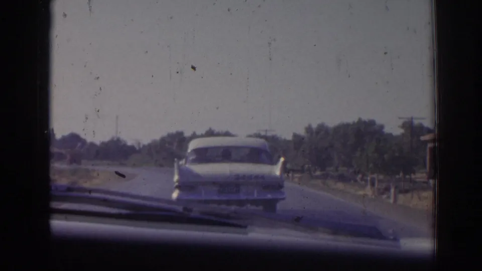 The white sedan continues along a paved rural road in the Izmir region, framed by the windshield and soft 8mm film edges.